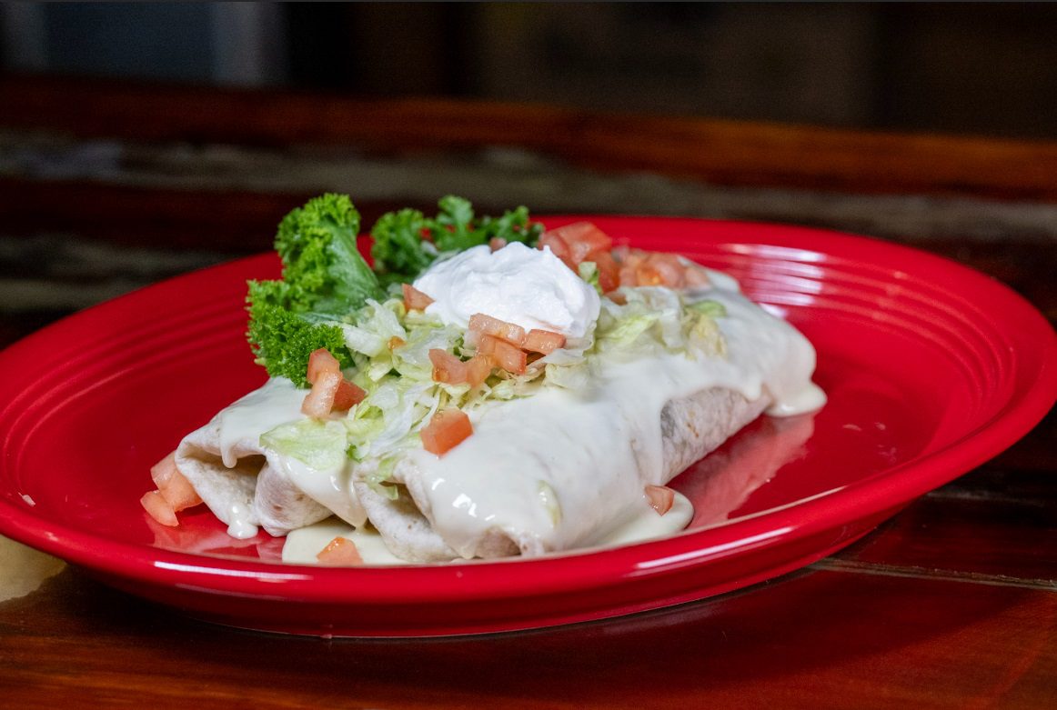Juan's Authentic Mexican Restaurant Mississippi - A burrito topped with melted white cheese sauce, shredded lettuce, diced tomatoes, sour cream, and a sprig of parsley—just the kind of flavor you'd find at a great family Mexican restaurant in Poplarville MS. Served on a red plate on a wooden table.