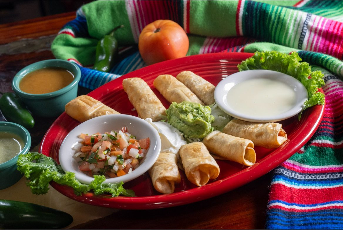 Juan's Authentic Mexican Restaurant Mississippi - A red plate with six taquitos, served with bowls of white cheese sauce, guacamole, sour cream, and pico de gallo, garnished with lettuce—an inviting spread from a Family Mexican restaurant Poplarville MS. Colorful textiles and an orange tomato in the background.