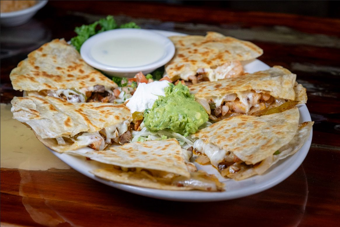 Juan's Authentic Mexican Restaurant Mississippi - A plate of quesadillas cut into wedges, filled with cheese and meat. In the center are guacamole, sour cream, and a small bowl of white dipping sauce—just what you’d expect from the best tacos near me Gulf Coast MS—garnished with lettuce and pico de gallo.