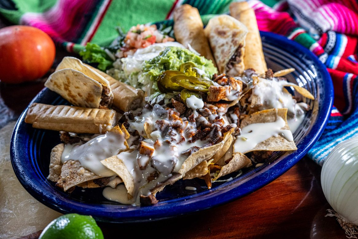 Juan's Authentic Mexican Restaurant Mississippi - A blue plate filled with nachos topped with grilled meat, cheese, jalapeños, and sour cream, served with guacamole, pico de gallo, lettuce, and taquitos on a colorful cloth at a Family Mexican restaurant Poplarville MS.