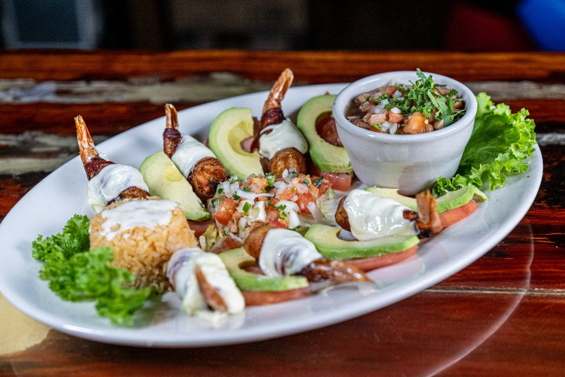 Juan's Authentic Mexican Restaurant Mississippi - A white plate with bacon-wrapped shrimp drizzled in white sauce, avocado and tomato slices, rice, lettuce, and fresh salsa—just one of the tempting dishes at our Mexican restaurant in Long Beach MS.