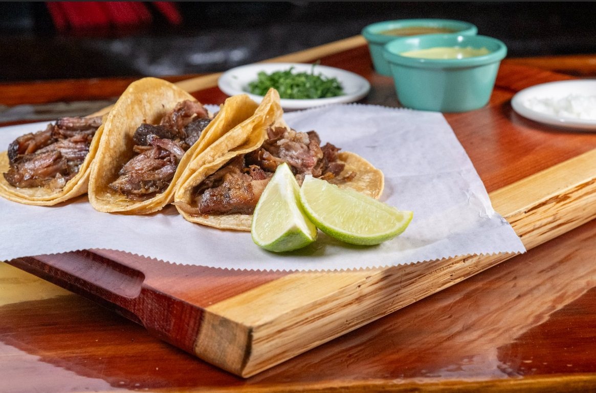 Juan's Authentic Mexican Restaurant Mississippi - Three beef tacos with corn tortillas are served on parchment paper with lime wedges. In the background, small dishes of chopped herbs, creamy sauce, and sliced onions evoke the flavors of authentic Mexican food Biloxi Mississippi.